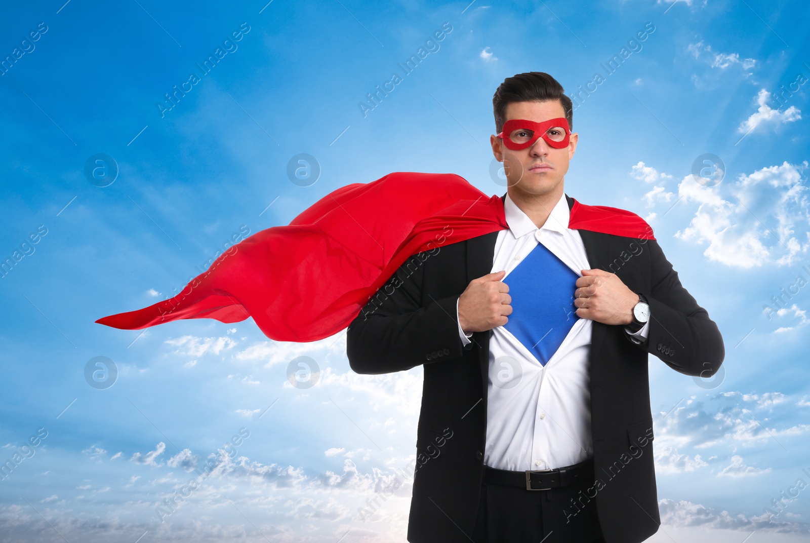 Image of Confident man wearing superhero cape and mask taking suit off against blue sky with clouds