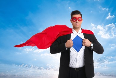 Image of Confident man wearing superhero cape and mask taking suit off against blue sky with clouds