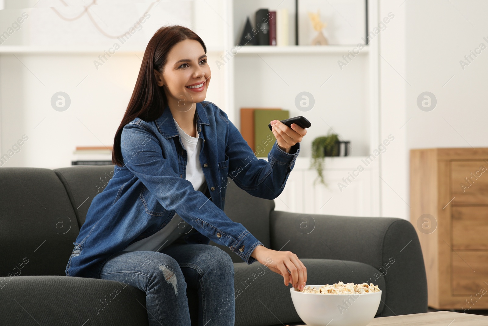 Photo of Happy woman changing TV channels with remote control on sofa at home