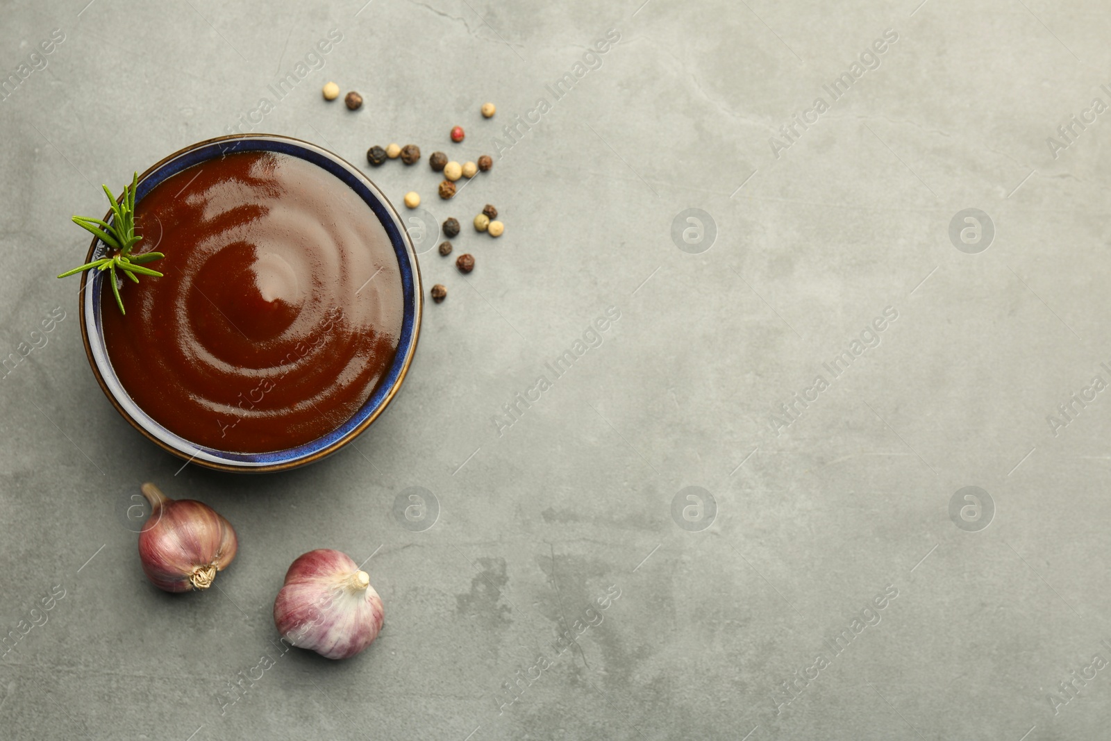 Photo of Tasty barbeque sauce in bowl, rosemary, garlic and peppercorns on grey textured table, flat lay. Space for text
