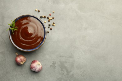 Photo of Tasty barbeque sauce in bowl, rosemary, garlic and peppercorns on grey textured table, flat lay. Space for text