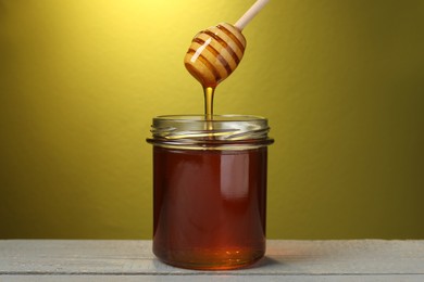 Photo of Pouring tasty honey from dipper into glass jar on grey wooden table against golden background