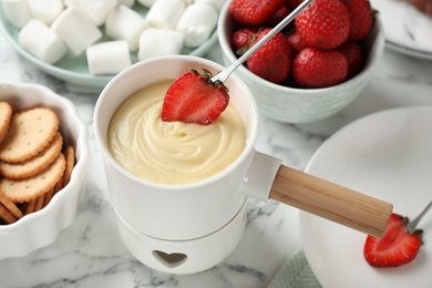 Photo of Dipping strawberry into fondue pot with white chocolate on marble table