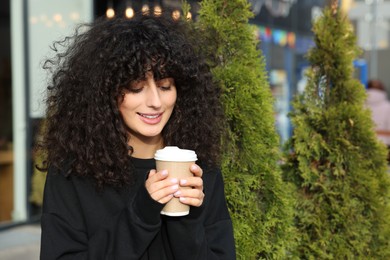 Happy young woman in stylish black sweater with cup of coffee outdoors, space for text