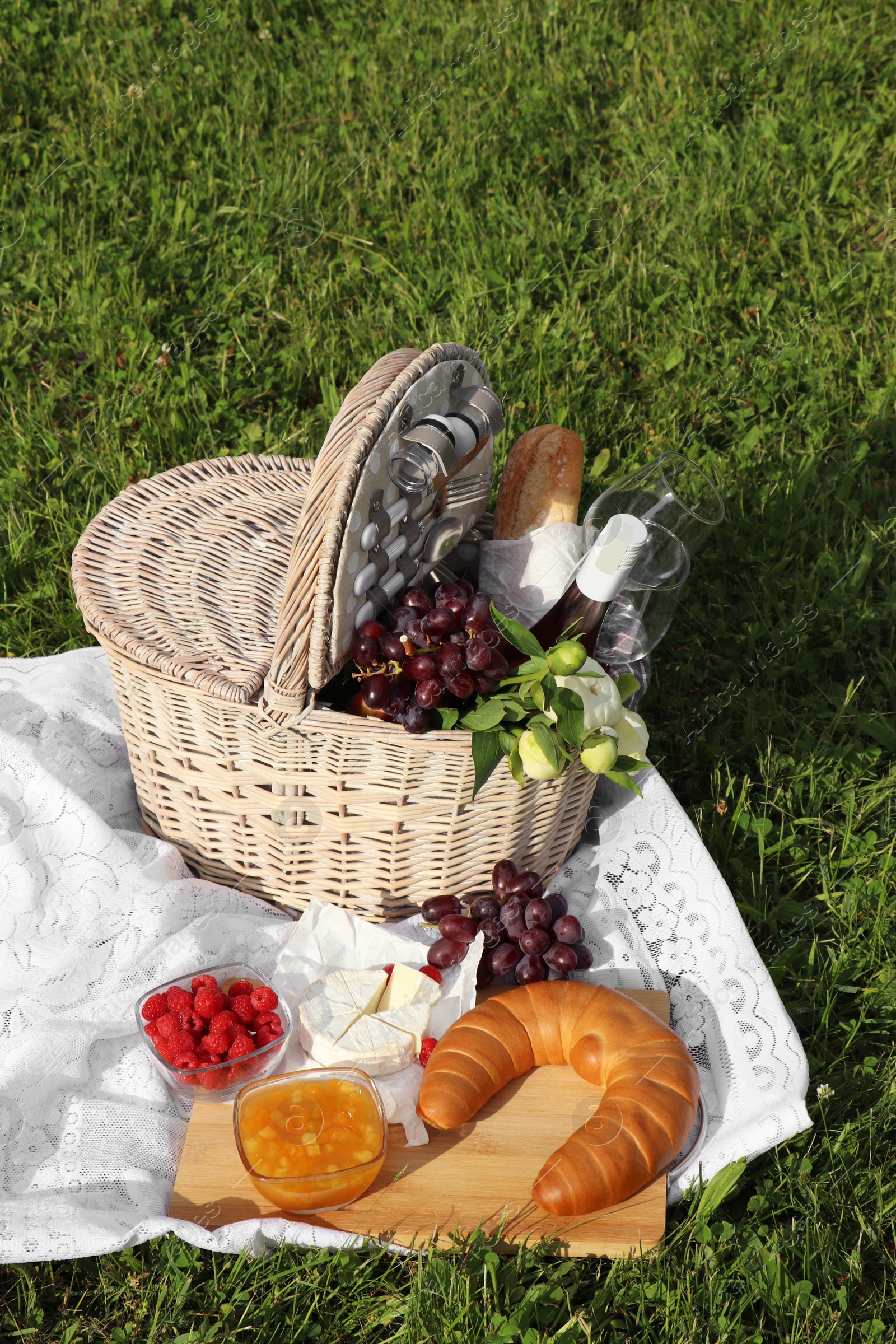 Photo of Picnic blanket with tasty food, flowers, basket and cider on green grass outdoors