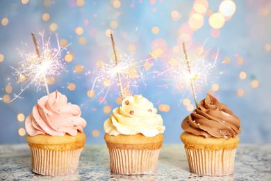 Image of Delicious birthday cupcakes with sparklers on table against blurred lights