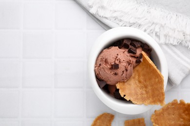 Tasty chocolate ice cream and pieces of waffle cone in bowl on white tiled table, flat lay. Space for text