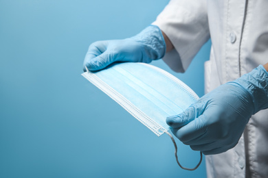 Doctor in latex gloves holding disposable face mask on light blue background, closeup. Protective measures during coronavirus quarantine