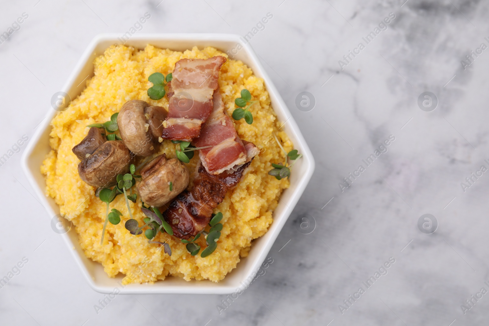 Photo of Cooked cornmeal with bacon, mushrooms and microgreens in bowl on white marble table, top view. Space for text