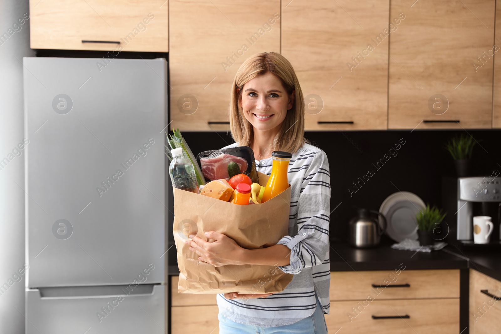 Photo of Woman with paper bag full of products near refrigerator in kitchen
