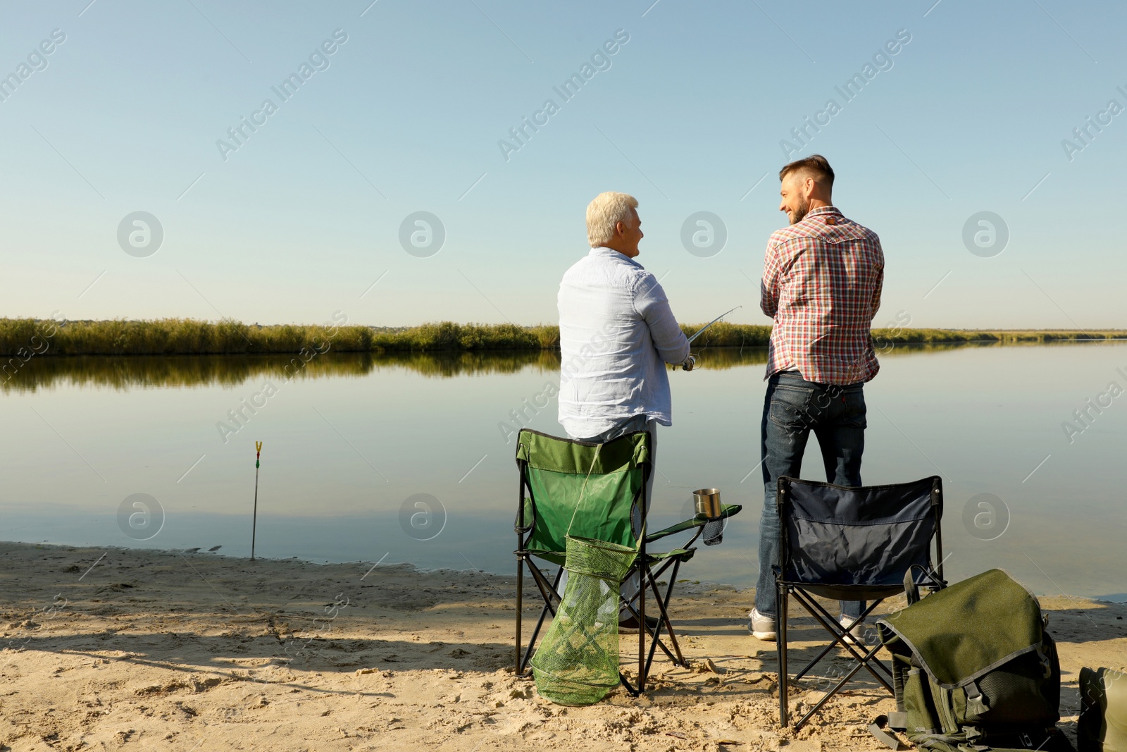 Photo of Father and adult son fishing together from riverside on sunny day