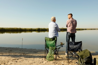 Photo of Father and adult son fishing together from riverside on sunny day