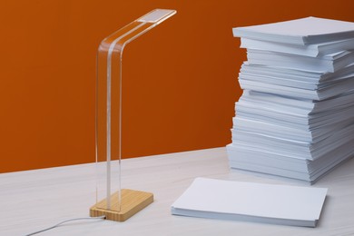 Stack of paper sheets and lamp on white wooden table near orange wall