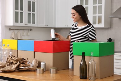 Photo of Garbage sorting. Smiling woman throwing metal can into cardboard box at table in kitchen