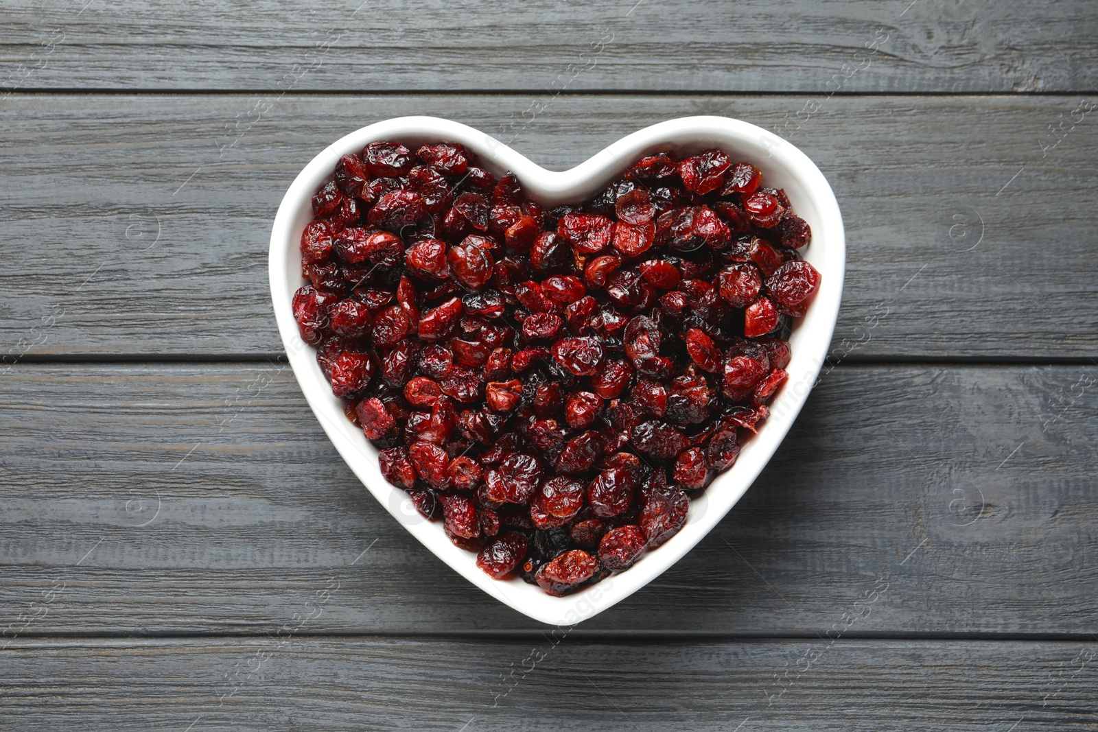 Photo of Heart shaped bowl with cranberries on wooden background, top view. Dried fruit as healthy snack