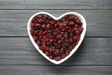 Photo of Heart shaped bowl with cranberries on wooden background, top view. Dried fruit as healthy snack