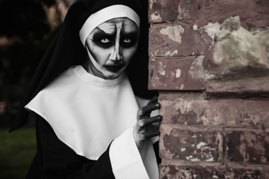 Scary devilish nun hiding behind red brick wall outdoors. Halloween party look