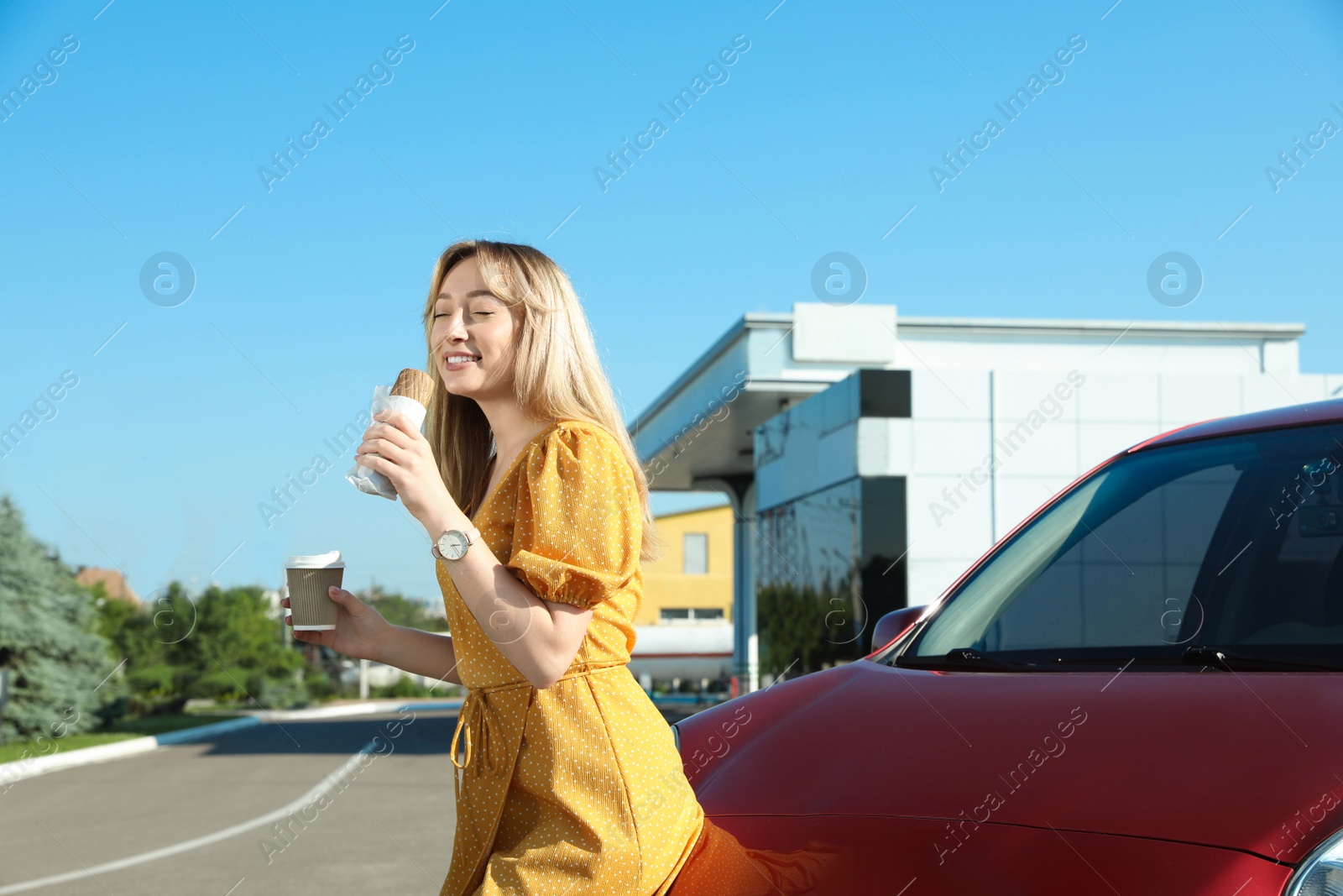 Photo of Beautiful young woman with coffee eating hot dog near car at gas station