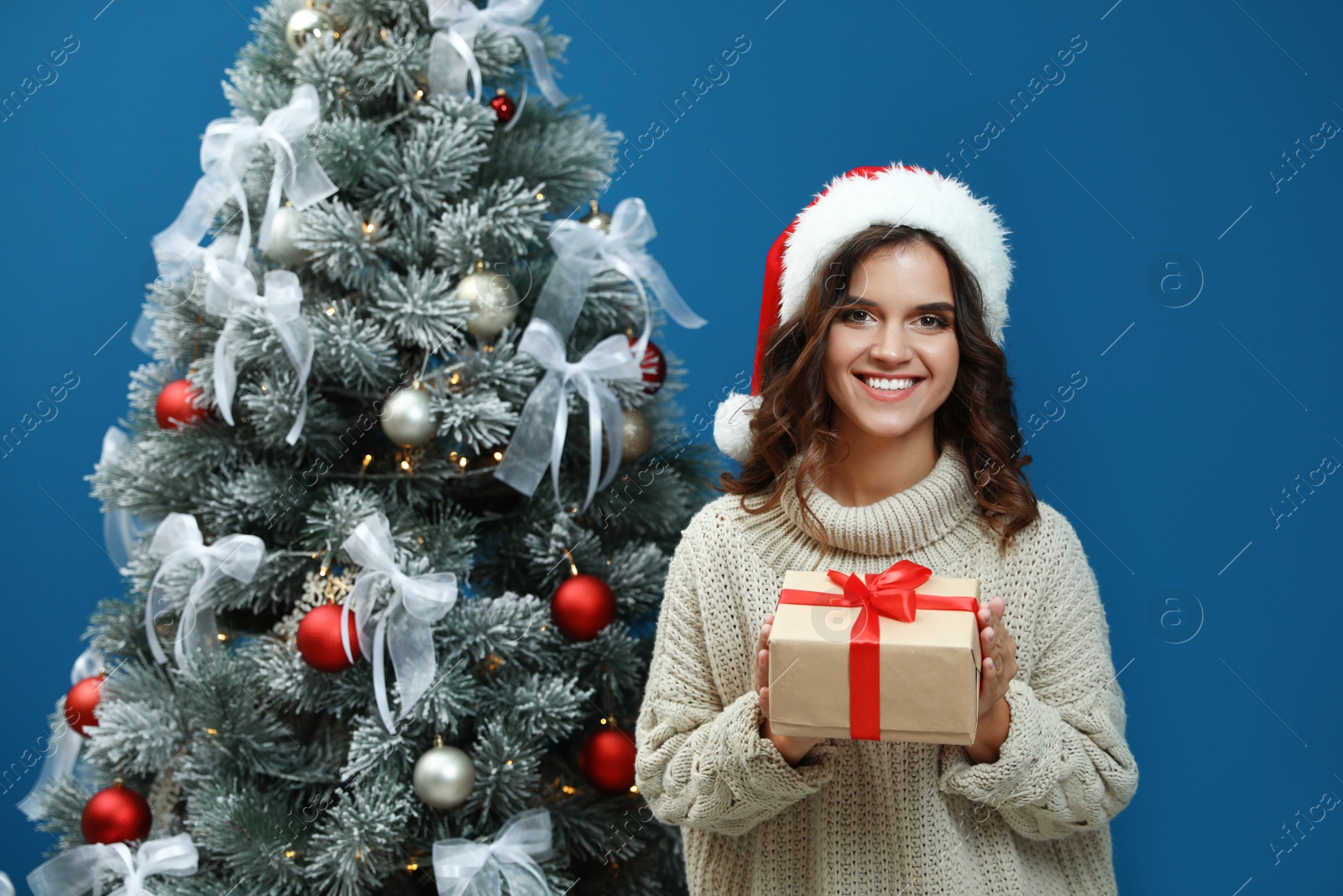 Image of Happy young woman in Santa hat with gift near Christmas tree on blue background