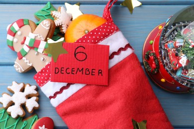 December, 6 - Saint Nicholas Day. Stocking with sweets, paper tag and festive decor on blue wooden table, flat lay