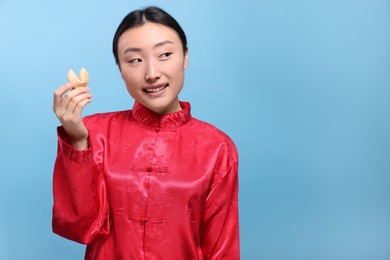 Photo of Asian woman holding tasty fortune cookie with prediction on light blue background, space for text