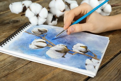 Photo of Woman painting cotton flowers in sketchbook at wooden table, closeup