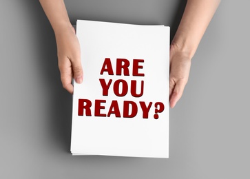 Image of Woman holding paper sheet with text Are You Ready? at grey table, top view