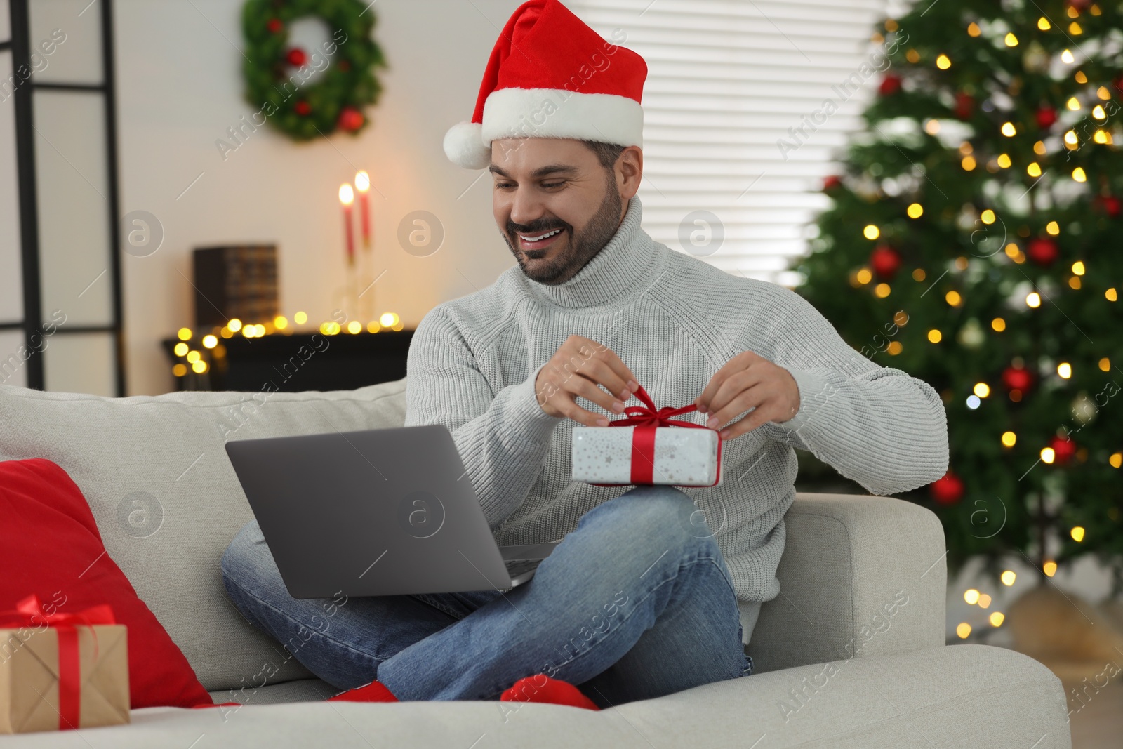 Photo of Celebrating Christmas online with exchanged by mail presents. Man in Santa hat opening gift box during video call on laptop at home