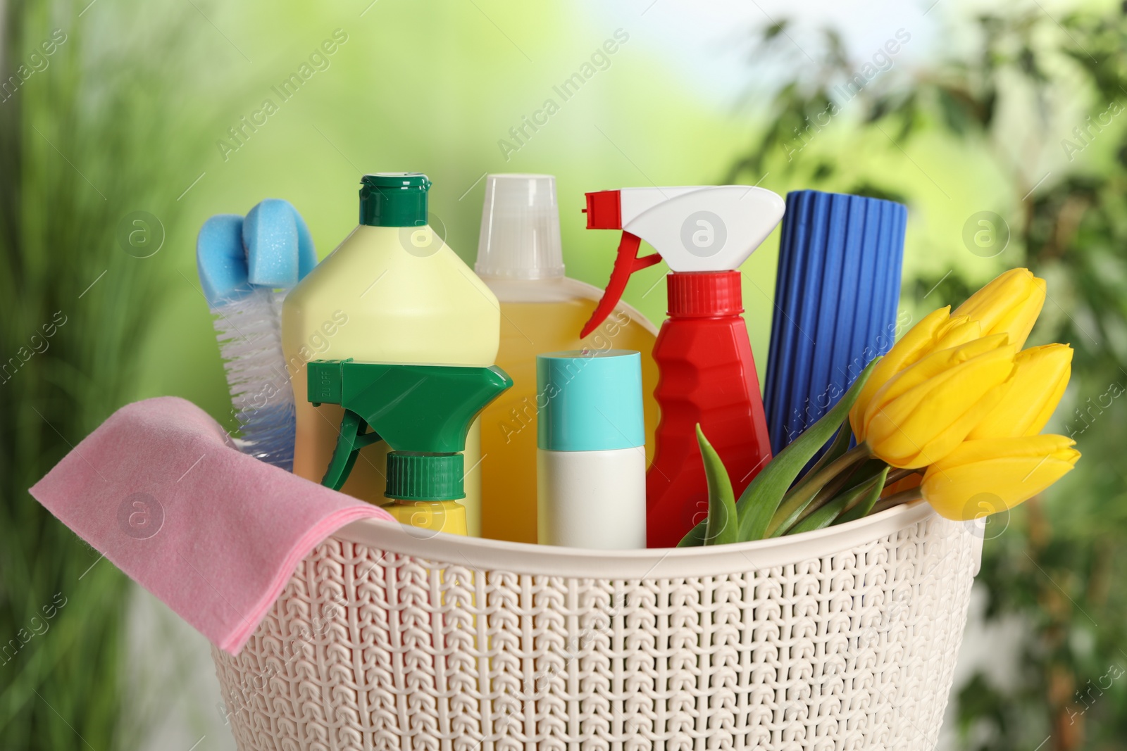 Photo of Spring cleaning. Plastic basket with detergents, supplies and beautiful flowers outdoors