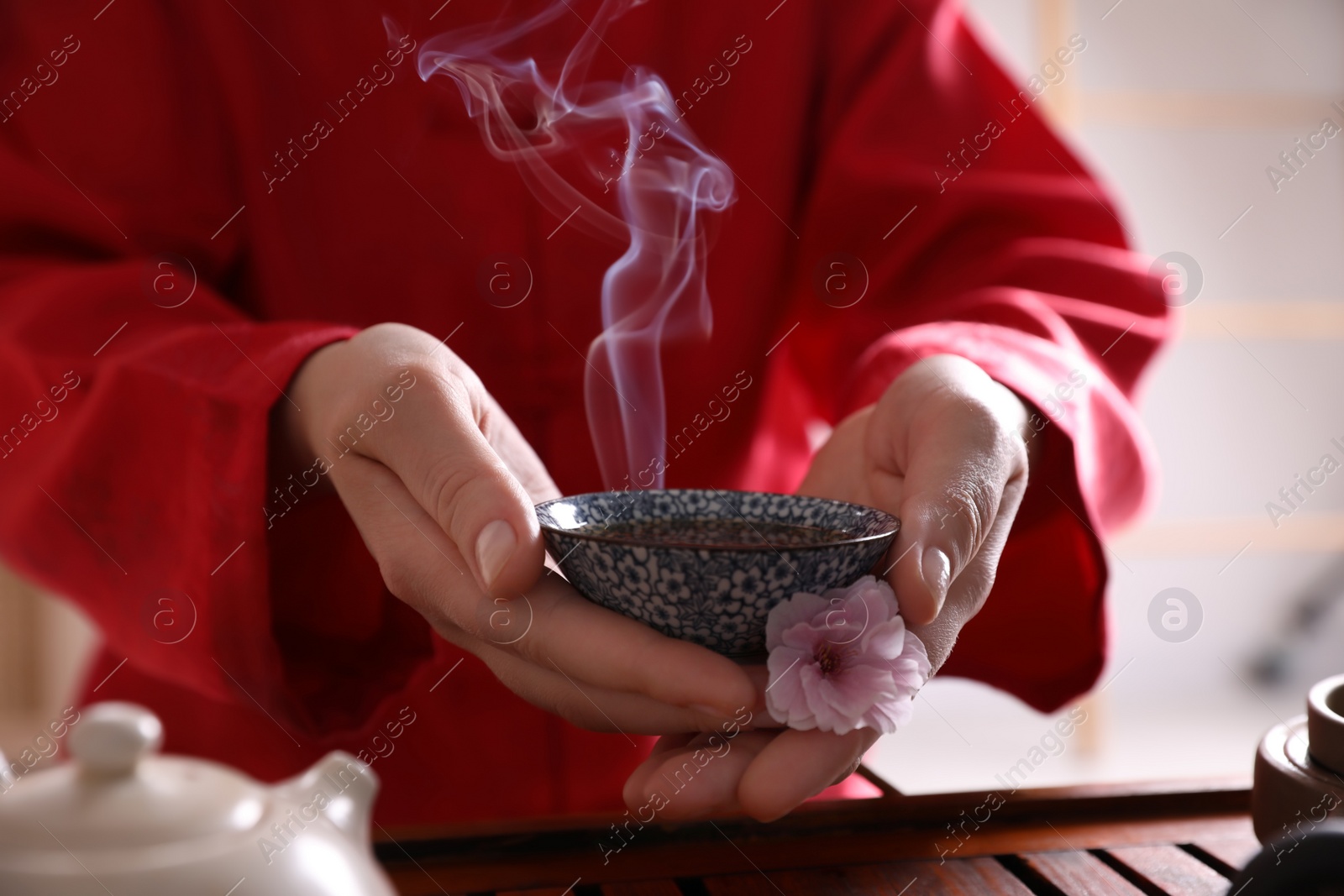 Photo of Master holding cup of freshly brewed tea and flower during traditional ceremony at table, closeup