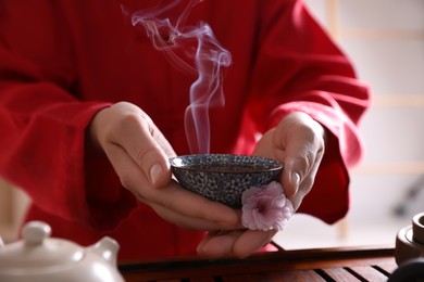 Photo of Master holding cup of freshly brewed tea and flower during traditional ceremony at table, closeup