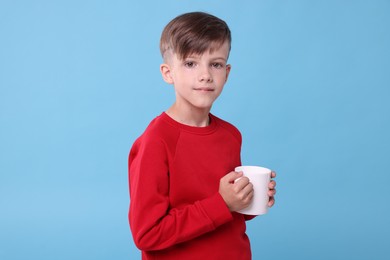 Cute boy with white ceramic mug on light blue background