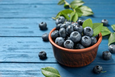 Photo of Tasty fresh blueberries with leaves in bowl on blue wooden table. Space for text