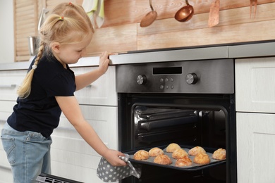 Photo of Little girl taking out baking sheet with cookies from oven in kitchen