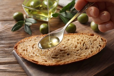 Photo of Woman pouring olive oil into spoon over bread at wooden table, closeup