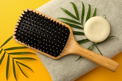 Wooden hairbrush, solid shampoo, towel and green leaves on orange background, flat lay