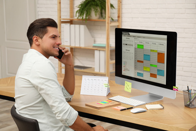Photo of Man talking on smartphone while working with calendar app at table in office