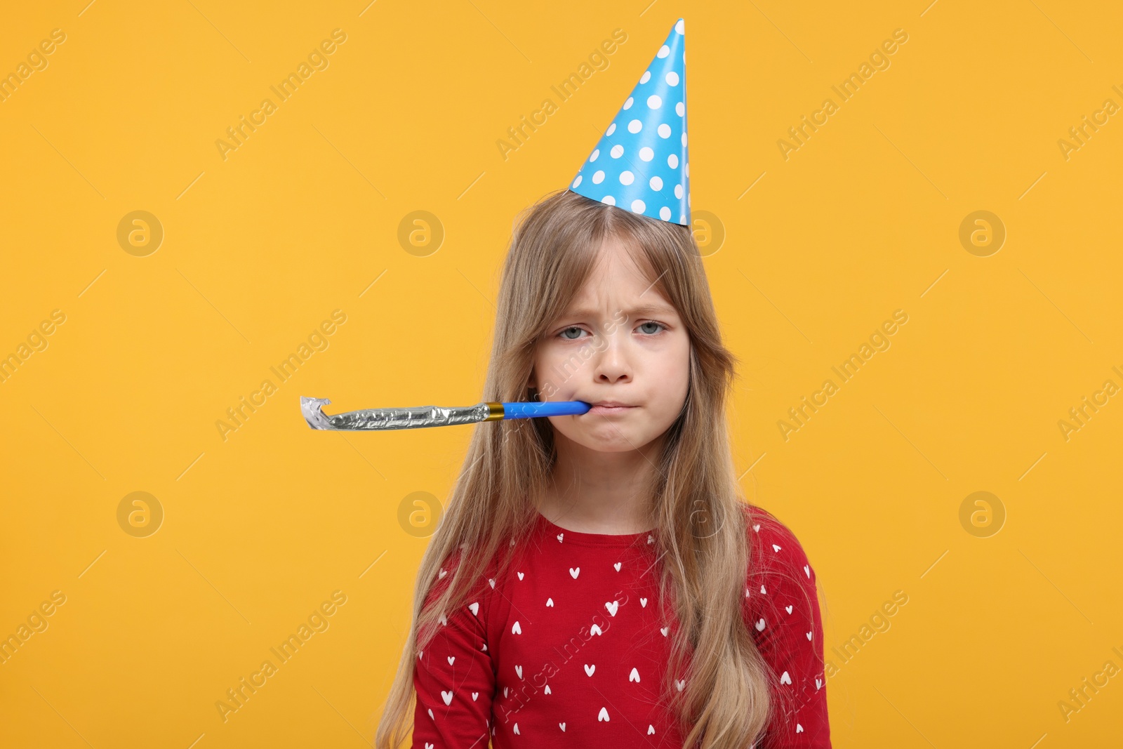 Photo of Unhappy little girl in party hat with blower on yellow background