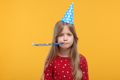 Photo of Unhappy little girl in party hat with blower on yellow background