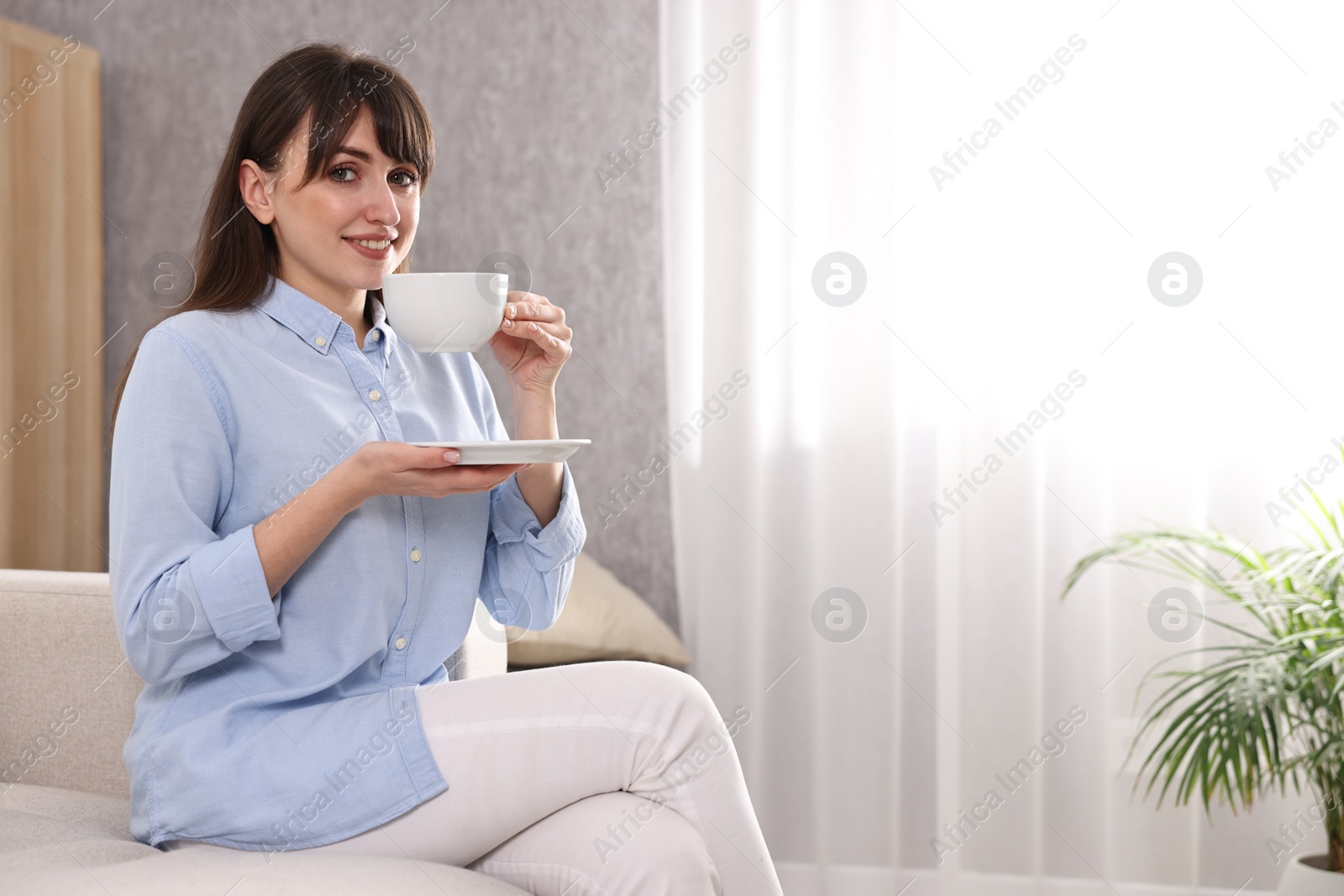 Photo of Beautiful young housewife with cup of drink on sofa at home, space for text