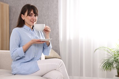 Beautiful young housewife with cup of drink on sofa at home, space for text