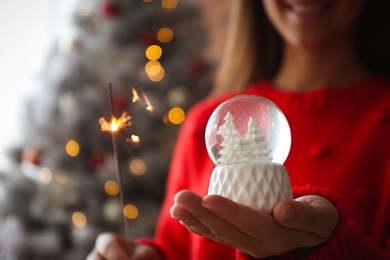 Woman with snow globe and sparkler near Christmas tree, closeup. Space for text