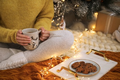 Woman with cup of hot drink and cookies sitting near Christmas tree on floor in room, closeup