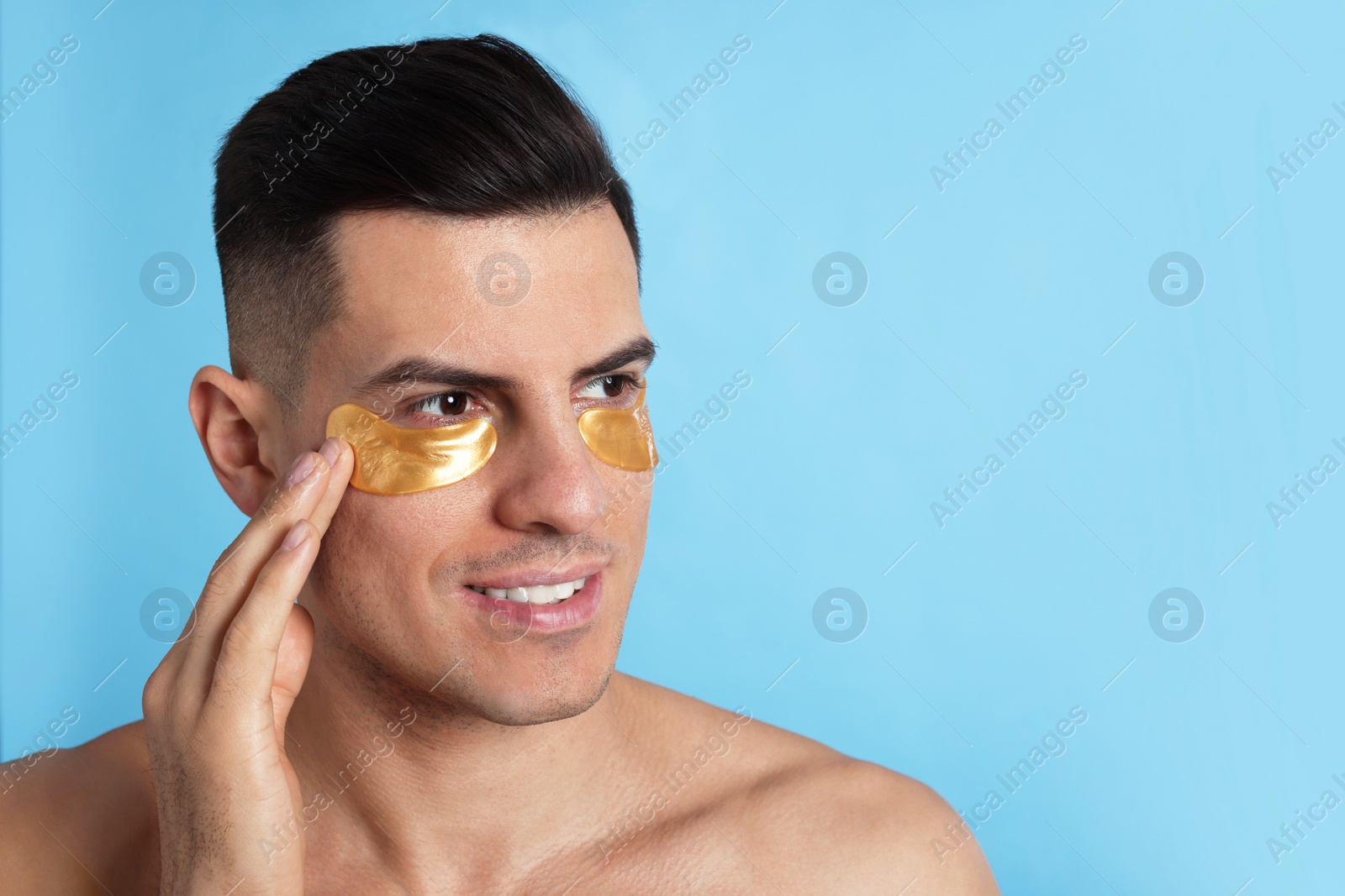 Photo of Man applying golden under eye patch on light blue background. Space for text