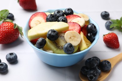 Photo of Tasty fruit salad in bowl and ingredients on white tiled table, closeup