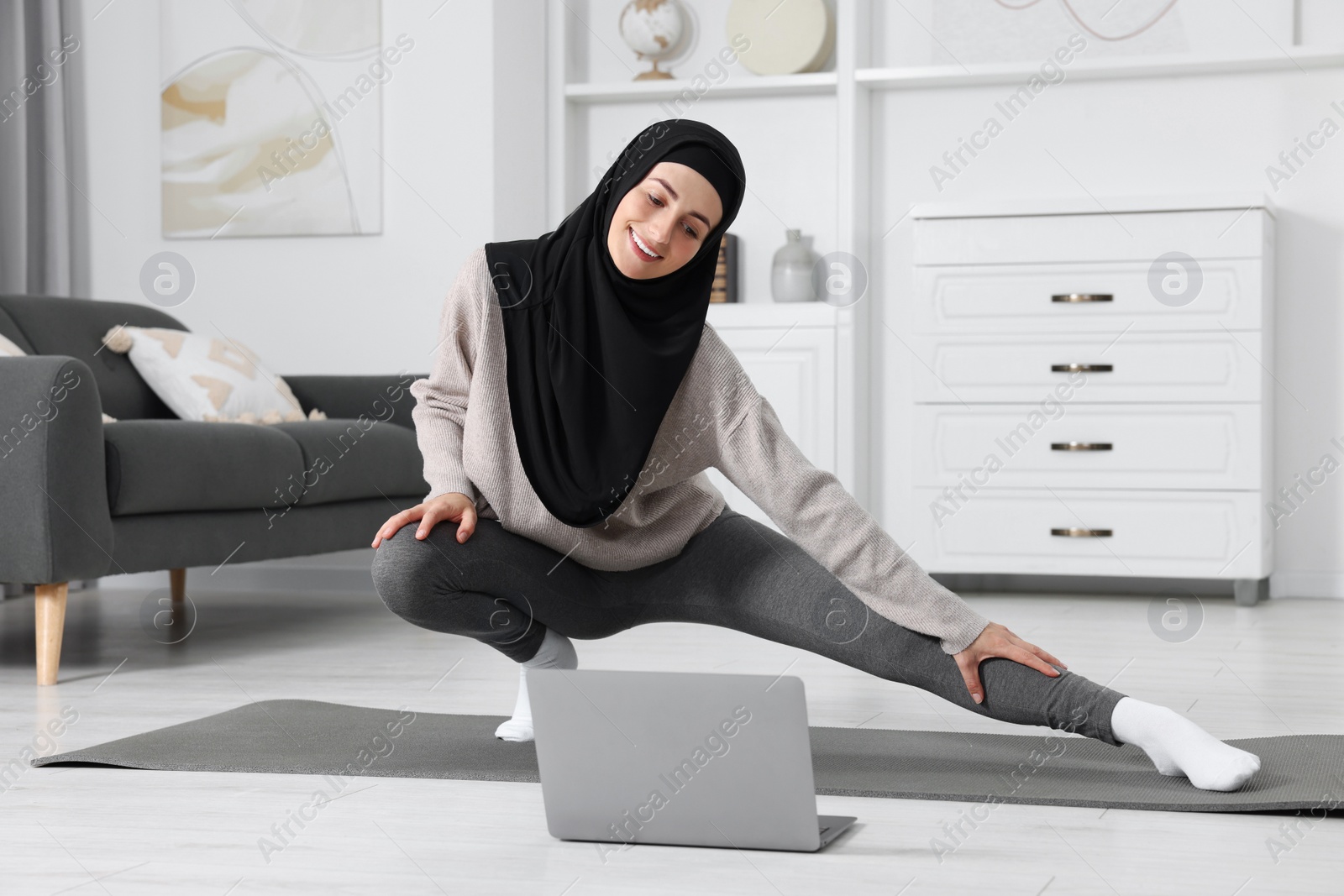 Photo of Muslim woman in hijab stretching near laptop on fitness mat at home