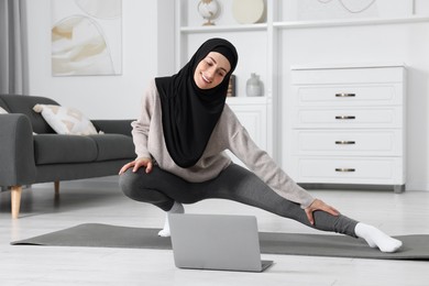 Photo of Muslim woman in hijab stretching near laptop on fitness mat at home