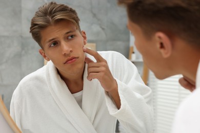 Photo of Upset young man looking at mirror and touching pimple on his face indoors. Acne problem