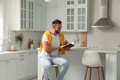 Photo of Handsome man reading book on stool in kitchen