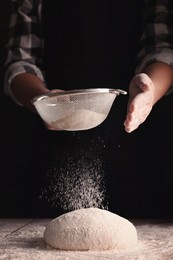 Man sprinkling flour over dough at wooden table on dark background, closeup